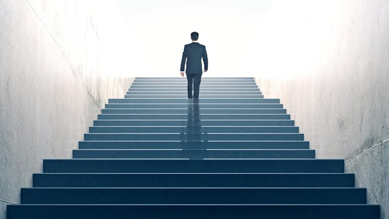 Suited businessperson walking up a multiflight stairway, a few steps from the top